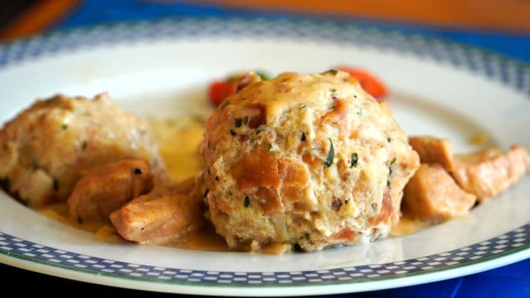 Nahaufnahme: Zwei große Semmelknödel und saftige Fleischstücke, großzügig bedeckt mit cremiger Soße, ansprechend serviert auf einem dekorativen Teller.