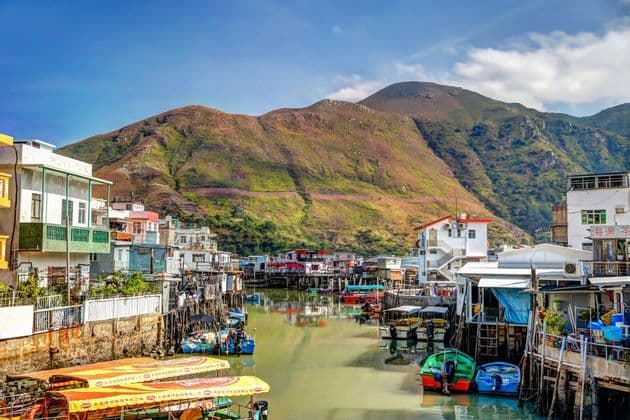 A colorful village of stilt houses and boats sits on a waterway at the base of a large, green mountain under a blue sky.