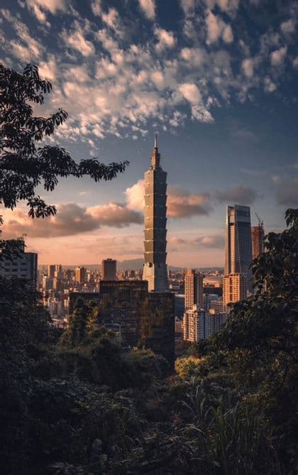 La torre Taipei 101 si erge sopra uno skyline cittadino, incorniciata da una fitta vegetazione scura, sotto un cielo parzialmente nuvoloso al tramonto.