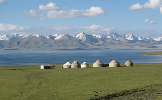 Eine Reihe Jurten steht auf einer Grasfläche neben einem großen See, mit einer Kette schneebedeckter Berge im Hintergrund.