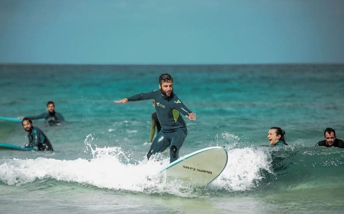 Un uomo del gruppo WeRoad, in muta, sta su una tavola da surf cavalcando un'onda, mentre altri osservano dall'oceano turchese.