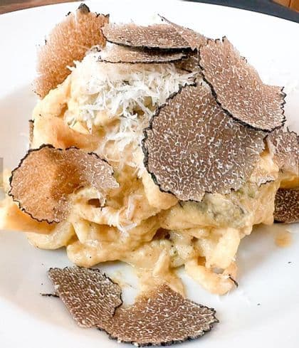 Primo piano di un piatto di pasta con salsa cremosa, guarnito con abbondanti scaglie di tartufo nero e formaggio grattugiato, servito su un piatto bianco.