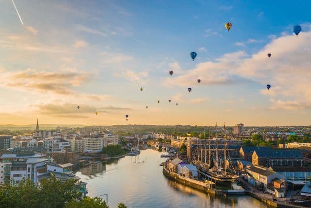 Numerose mongolfiere fluttuano sopra una città portuale con un veliero ormeggiato all'alba sotto un cielo parzialmente nuvoloso.