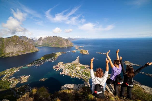 A WeRoad group trip of three women sitting on a clifftop, arms raised, overlooking a coastal village surrounded by mountains and sea.