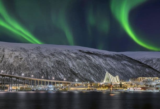 The green Northern Lights glow in the night sky over an illuminated city with a modern cathedral, next to a snowy mountain.