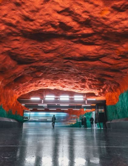Un train traverse une station de métro avec un plafond rouge évoquant une grotte et des murs peints, alors que des passagers attendent sur le quai réfléchissant.