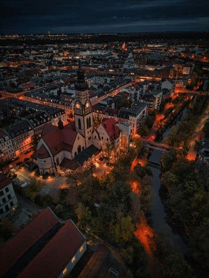 Eine Luftaufnahme einer Stadt bei Nacht, mit einer beleuchteten Kirche und einem Glockenturm im Vordergrund neben einem Fluss.