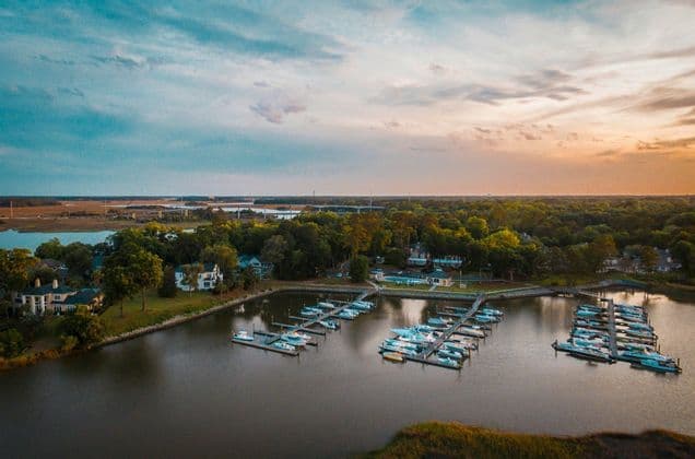 An aerial view of a marina with boats docked along piers, with a tree-lined shore and houses under a sunset sky.