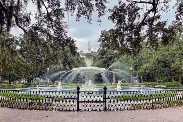 An ornate white tiered fountain with statues sprays water in a park, surrounded by trees and a black iron fence.