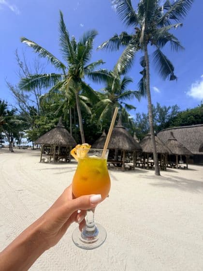 Une main tenant un cocktail tropical sur une plage de sable blanc avec des palmiers et des cabanes au toit de chaume en arrière-plan.