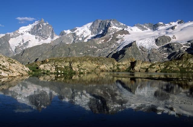 Une chaîne de montagnes enneigée se reflète parfaitement dans l'eau calme d'un lac alpin sous un ciel bleu clair.