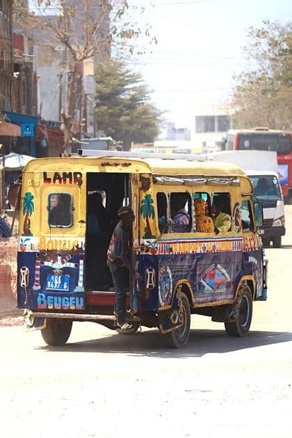 Ein Mann hängt sich an das Heck eines bunt bemalten Minibusses, der mit Fahrgästen eine Stadtstraße entlangfährt.