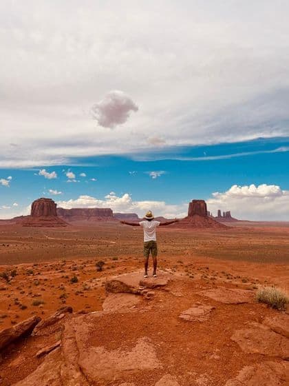 A person in a hat stands with arms outstretched on a rocky ledge, overlooking a vast desert landscape with large rock formations.