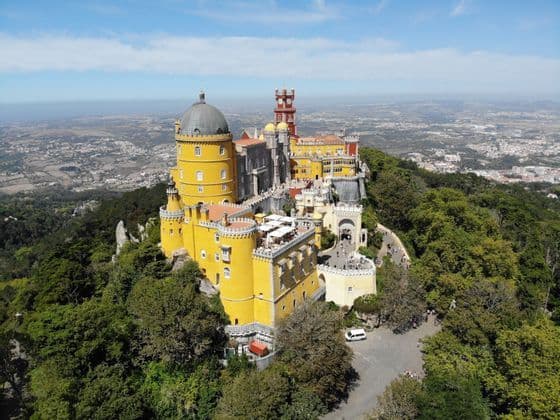 Vue aérienne d'un grand château jaune et rouge, perché sur une colline verdoyante et boisée, surplombant un vaste paysage urbain.