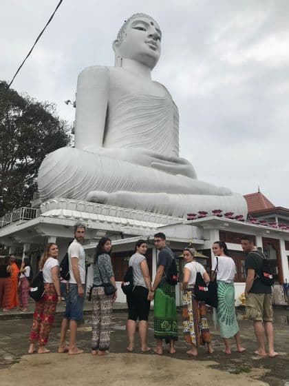 Eine WeRoad-Gruppenreise posiert barfuß vor einer riesigen weißen sitzenden Buddha-Statue in einem Tempel.