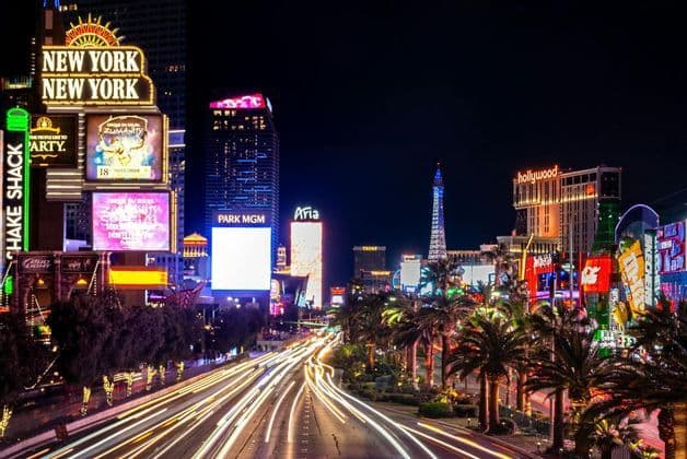 A long-exposure photograph of a busy city street at night, with traffic light trails and illuminated buildings with neon signs.