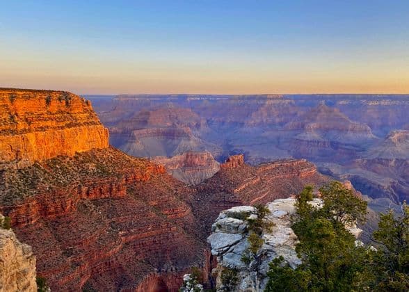 A vast, layered canyon with rock formations lit by warm sunrise light under a clear sky.