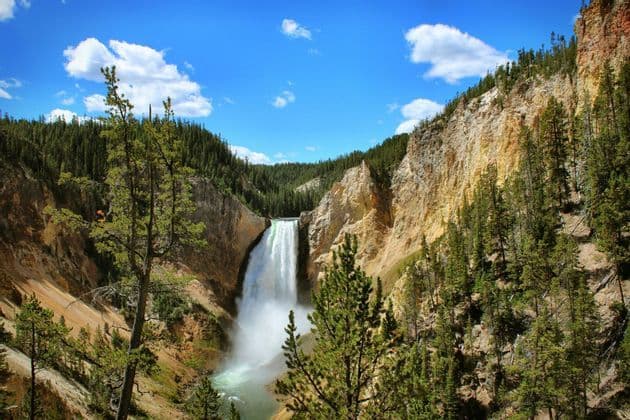 A large waterfall rushes into a river at the bottom of a deep, tree-lined canyon under a blue, partly cloudy sky.