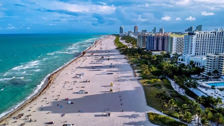 An aerial view of a wide sandy beach stretching along a city skyline, with turquoise ocean waves lapping the shore.