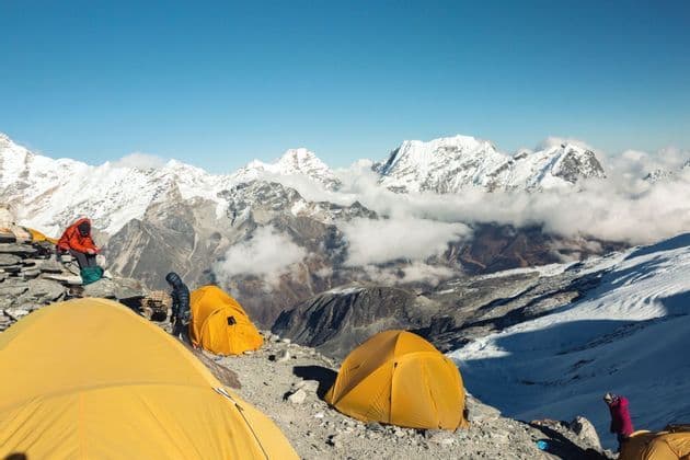 Un groupe WeRoad campe sous des tentes jaunes sur un flanc de montagne rocheux, avec une vaste chaîne de sommets enneigés en arrière-plan sous un ciel dégagé.