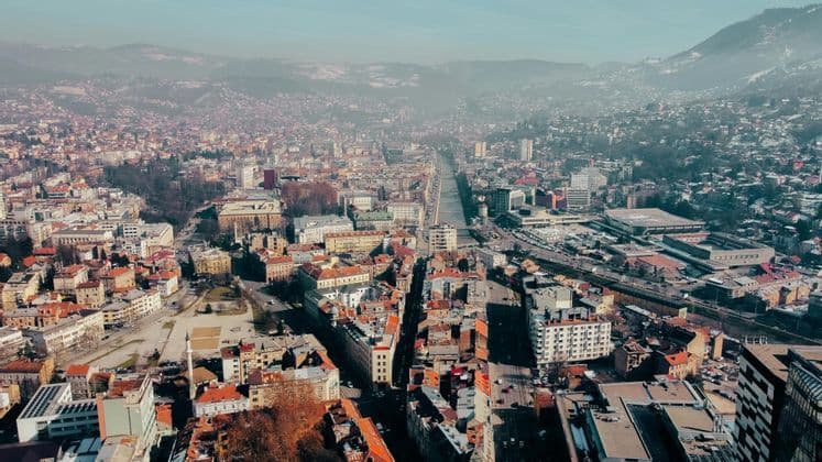 Una vista aerea di una città estesa con un fiume che la attraversa al centro, su uno sfondo di montagne nebbiose.