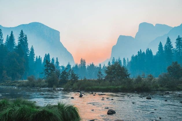 A rocky river flows through a pine forest valley with misty mountains in the background during a colorful sunset.