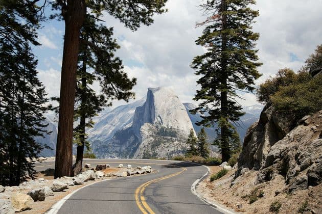 A winding paved road curves through a pine forest, with a view of a large granite dome and mountains under a partly cloudy sky.