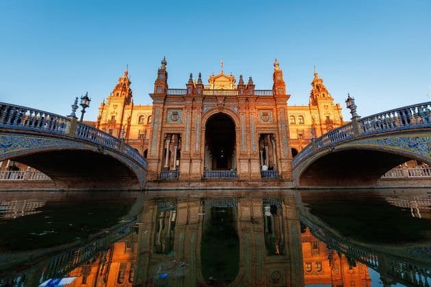 An ornate brick building with two tiled bridges is reflected in the water below during a clear sunset.