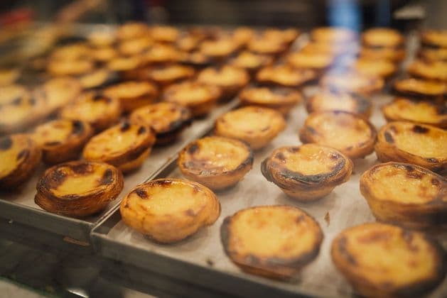Rows of freshly baked Portuguese custard tarts, pastéis de nata, on metal trays in a bakery display case.