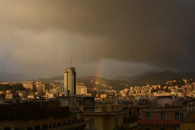A cityscape with a tall skyscraper under a dark, stormy sky, with a rainbow arching over distant hills.