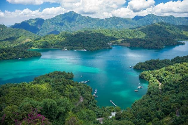 Vue aérienne d'un lac turquoise vif entouré de montagnes verdoyantes et boisées sous un ciel nuageux.