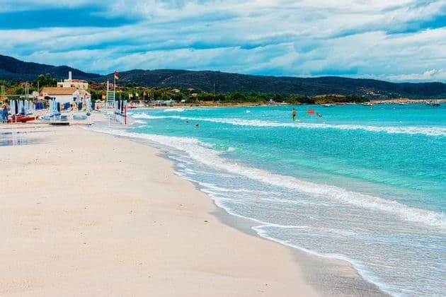 Una spiaggia di sabbia con acqua turchese che lambisce la riva, con ombrelloni e colline sotto un cielo nuvoloso.