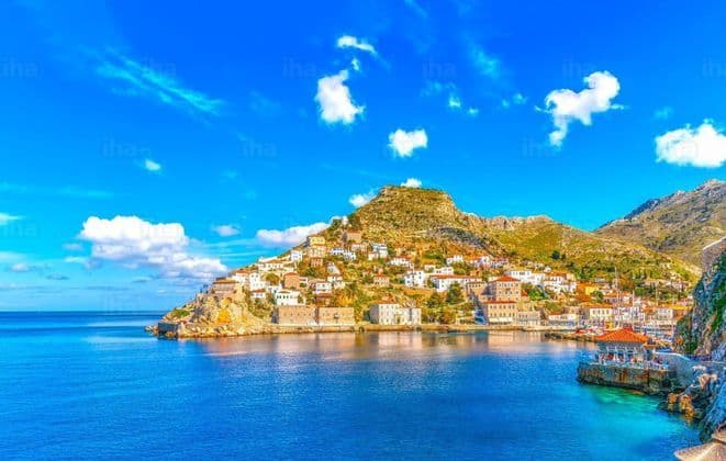 Una città costiera con case bianche e tetti in terracotta costruita su una collina che si affaccia su un mare blu calmo sotto un cielo soleggiato.