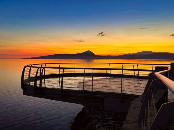 Una moderna piattaforma panoramica circolare si protende su un mare calmo, di fronte a montagne lontane sotto un cielo vibrante al tramonto.