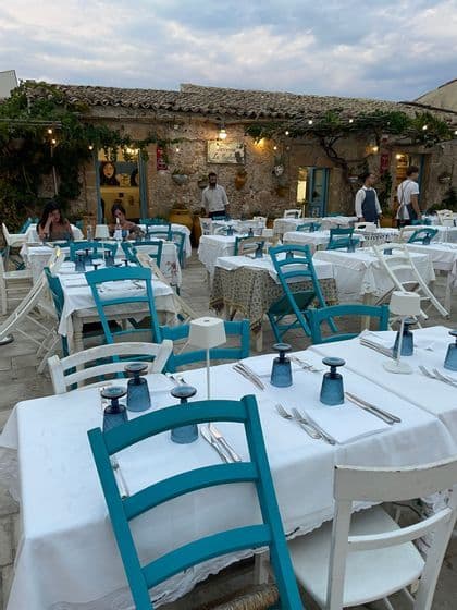 Tavoli apparecchiati con tovaglie bianche e sedie turchesi nel patio di un rustico ristorante in pietra al tramonto.