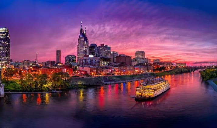 A wide-angle view of an illuminated city skyline next to a river during a vibrant purple and pink sunset, with a large riverboat on the water.