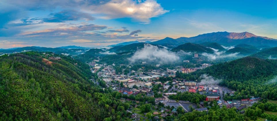 An aerial panoramic view of a town nestled in a lush, green mountain valley, with patches of clouds below a blue sky.