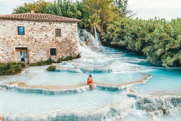Una donna in costume arancione siede sul bordo di una piscina termale a cascata accanto a un rustico edificio in pietra.