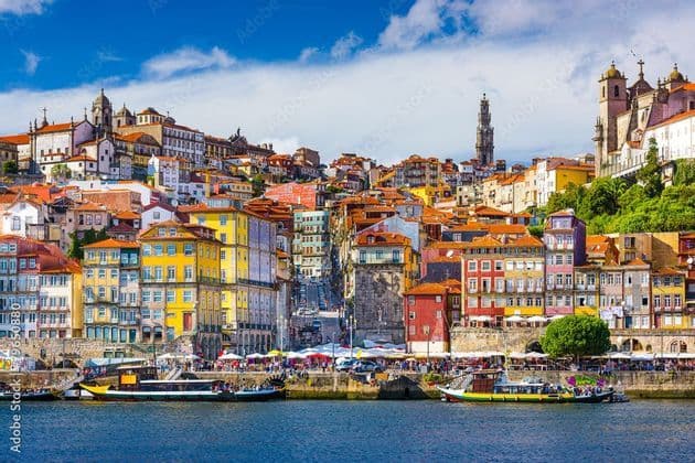 Colorful buildings of a hillside city seen from across a river, with boats docked along the waterfront under a blue sky.
