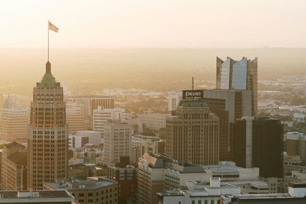 An aerial view of a cityscape with skyscrapers during a hazy sunset, with an American flag flying on a tall building.