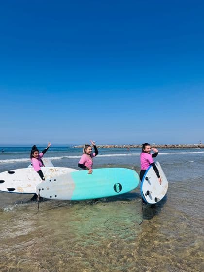 Tre donne in viaggio di gruppo WeRoad, sorridenti e in posa con le loro tavole da surf, mentre sono in piedi nell'oceano poco profondo.