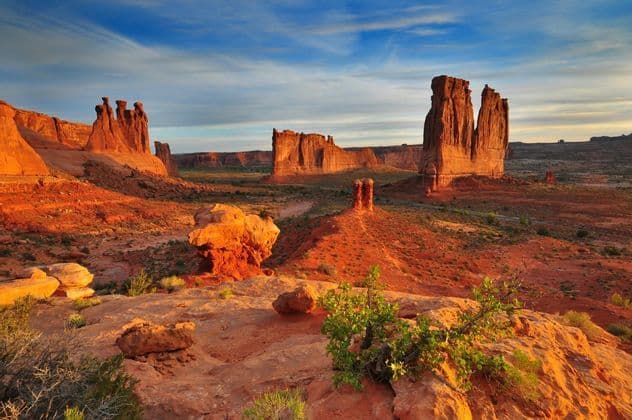 Red rock formations rise from a desert landscape, illuminated by warm sunrise light under a blue, partly cloudy sky.