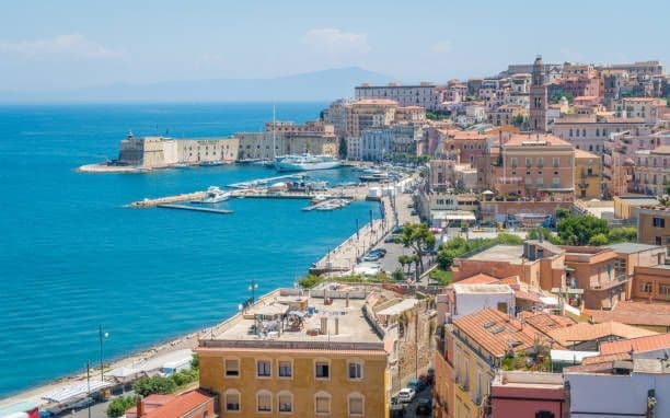 Veduta dall'alto di una cittadina costiera con edifici colorati, un porto con barche e una fortezza in pietra sul mare blu.