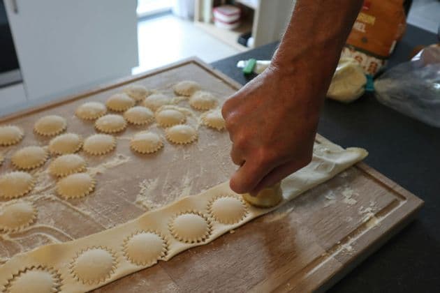 A person's hand uses a round cutter to make fresh ravioli from a sheet of pasta dough on a floured wooden board.