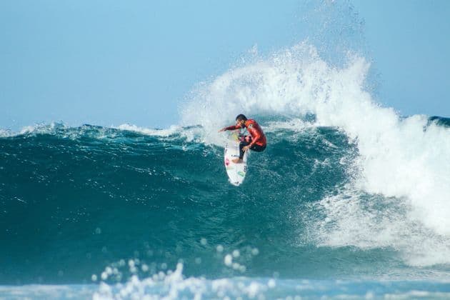 A surfer in a red wetsuit rides the crest of a large, breaking ocean wave under a clear blue sky.