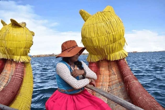 Una mujer con sombrero marrón y vestimenta tradicional colorida se sienta en un bote de totora con cabezas decorativas de gato, flotando sobre agua azul.