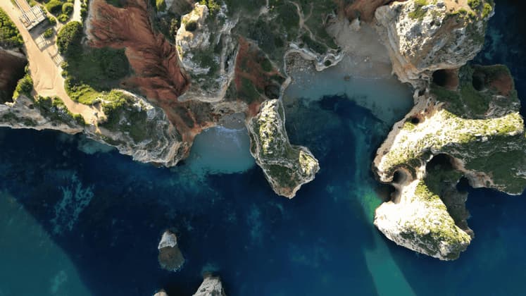 Una vista aerea, dall'alto, di una costa rocciosa con scogliere dalla cima verde e calette che lambiscono acque blu intenso e turchesi.