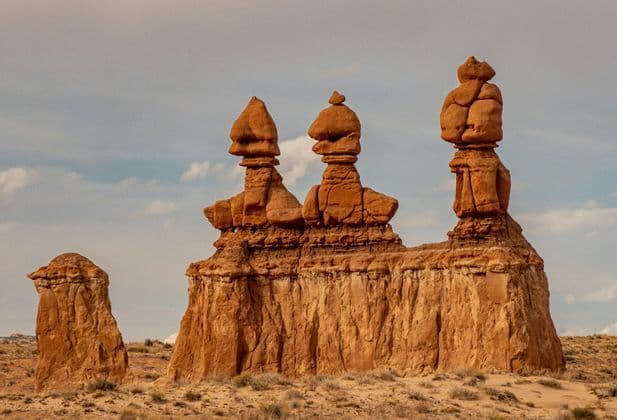 Three reddish-brown hoodoo rock formations stand on a ledge in a desert landscape under a partly cloudy sky.