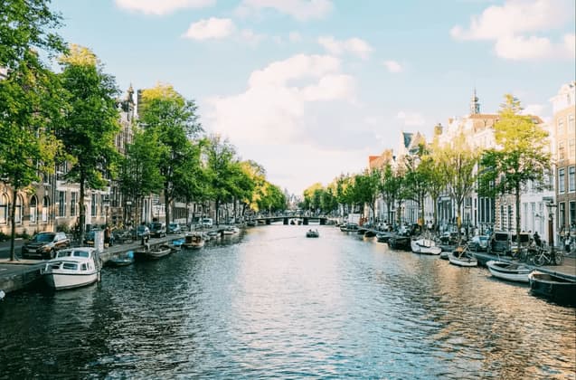 Una vista de un canal urbano con casas tradicionales, árboles verdes y barcos amarrados a sus orillas bajo un cielo azul con nubes blancas.