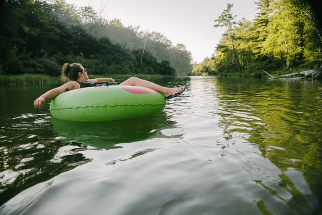 A woman relaxes in a green inflatable tube, floating down a calm river bordered by lush green trees.